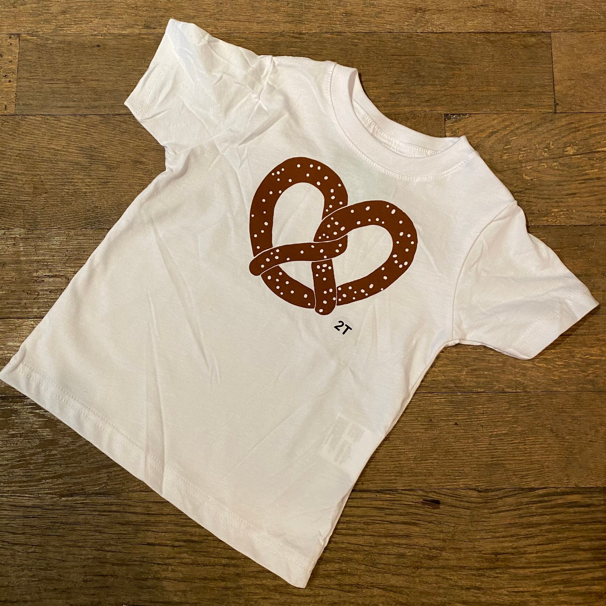 A white 100% cotton Pretzel Toddler T-Shirt with a Philly pretzel design by exit343design lying on a wooden floor.