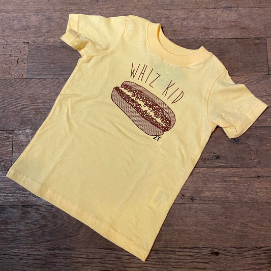 Yellow Whiz Kid toddler tee with hot dog graphic and "Philly genius" text displayed on a wooden floor by exit343design.